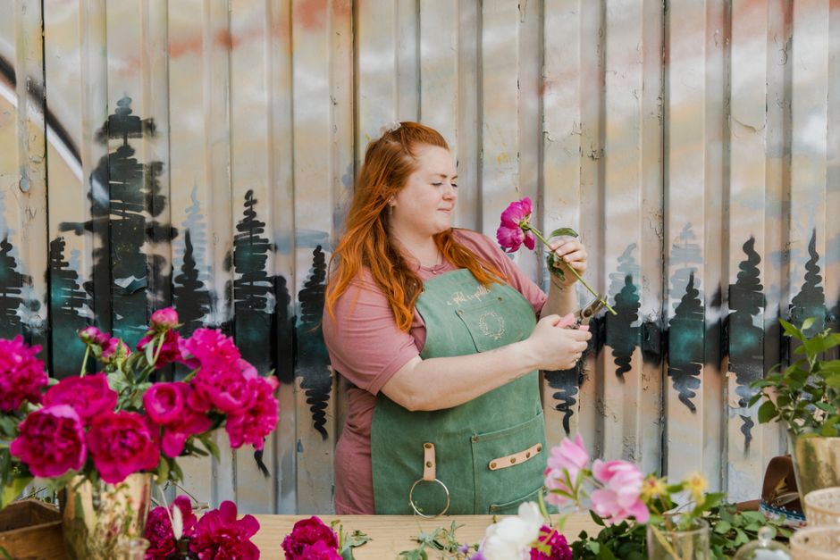 Redhead florist arranging pink peonies in an outdoor workshop, showcasing floral design skills.